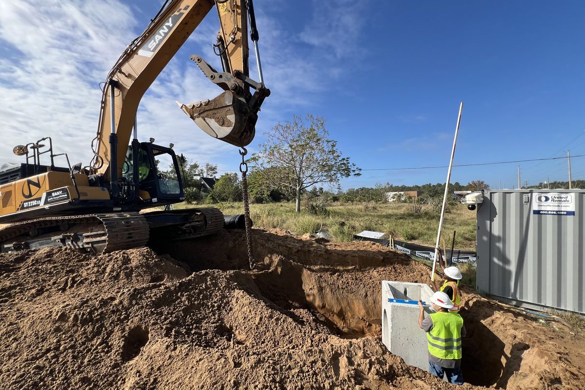 Excavator lowering a precast inlet into a trench with a crew member guiding it